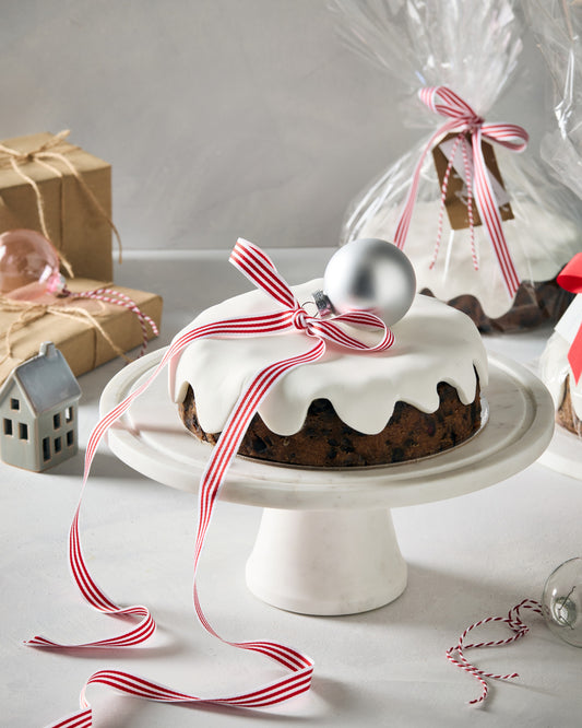 Christmas fruit cake with white fondant icing, a red and white striped ribbon and a silver bauble on a white cake stand
