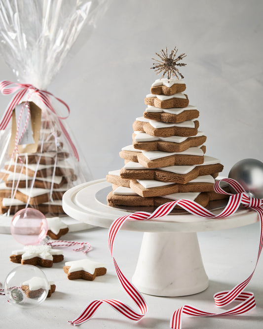 Gingerbread star tree of stacked iced star cookies on a white cake stand, with a gift wrapped star tree behind